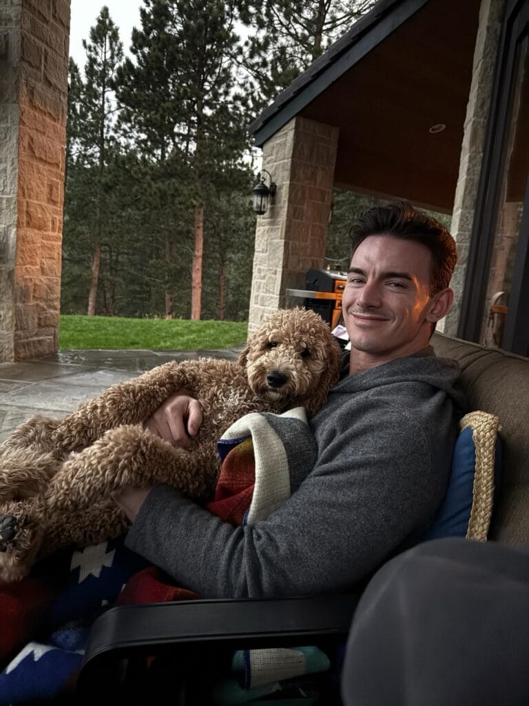 Noah Berkson relaxing on a covered stone patio holding a goldendoodle, with pine trees behind