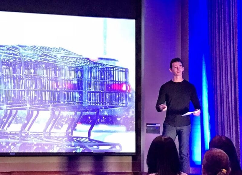 Noah Berkson speaking on stage next to a large screen showing shopping carts, in front of an audience