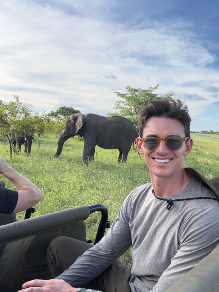 Noah Berkson smiling on a safari vehicle with elephants grazing in the grassland behind him